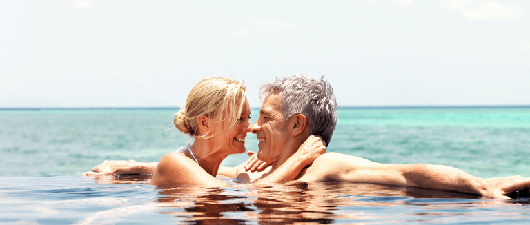 an attractive middle-aged couple, smiling and looking at one another, bathes in an infinity-edge pool overlooking the gulf behind it